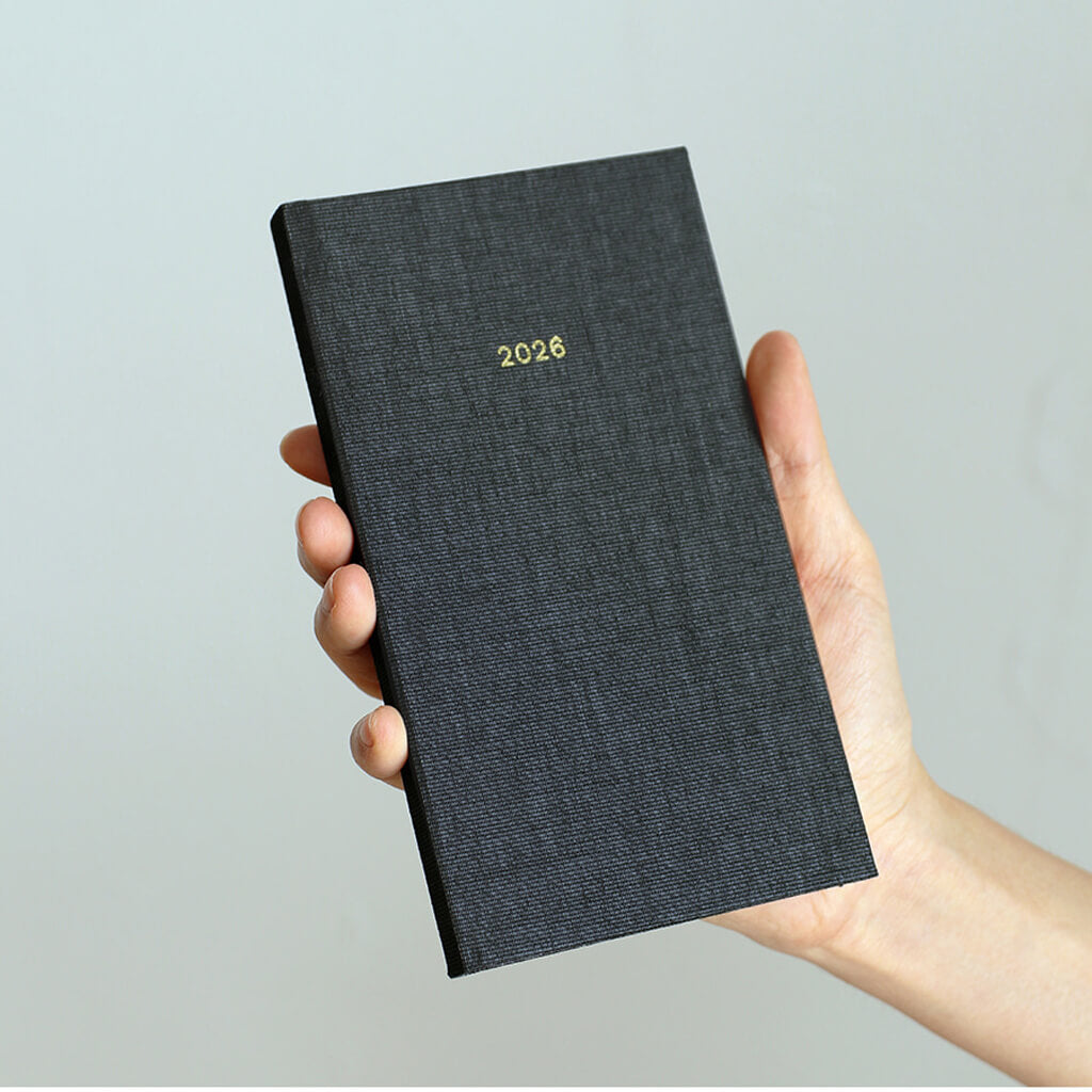 Hand holding a black textured diary with '2026' on a light gray background