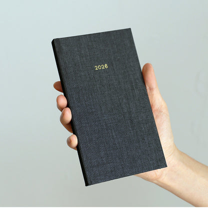 Hand holding a black textured diary with '2026' on a light gray background