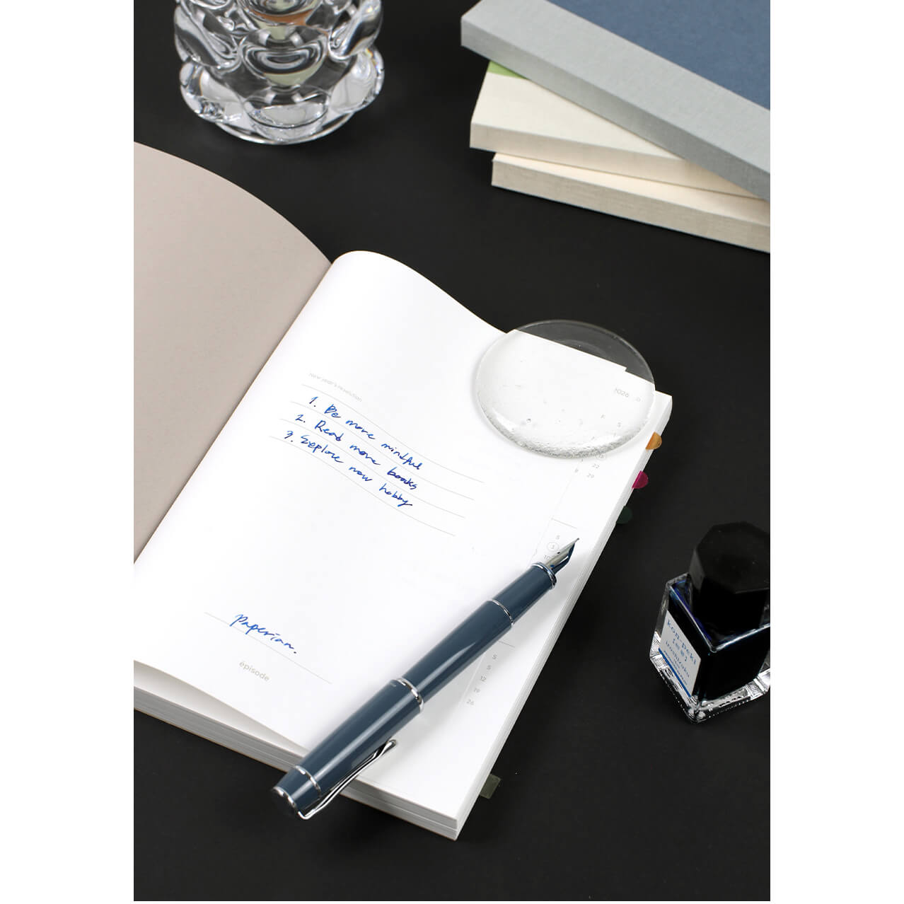 Fountain pen on an open notebook with ink bottle and glass container in the background