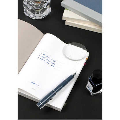 Fountain pen on an open notebook with ink bottle and glass container in the background