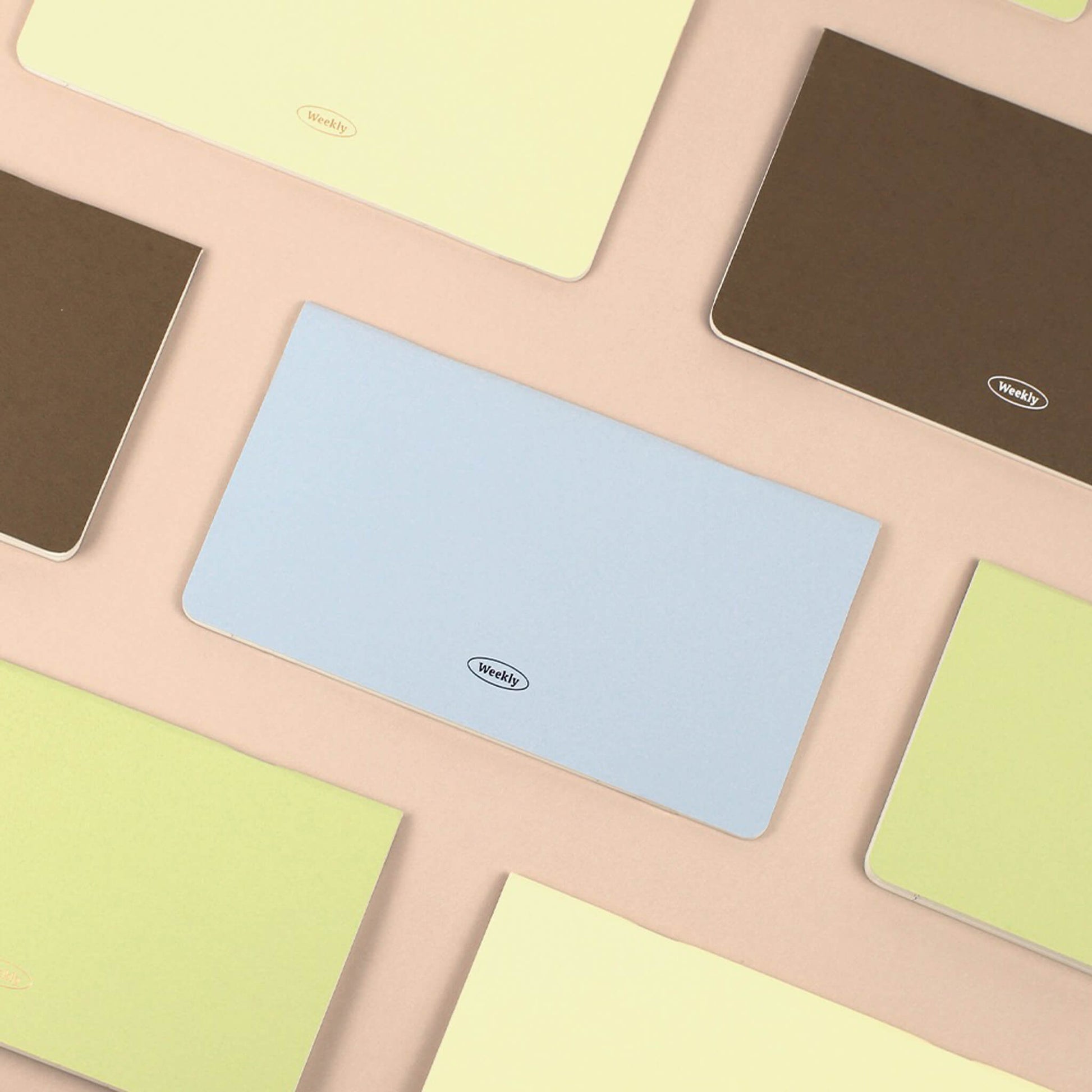 Colorful weekly planner with a brand logo on a patterned surface. The colors include baby blue, caramel brown, light yellow and light green.